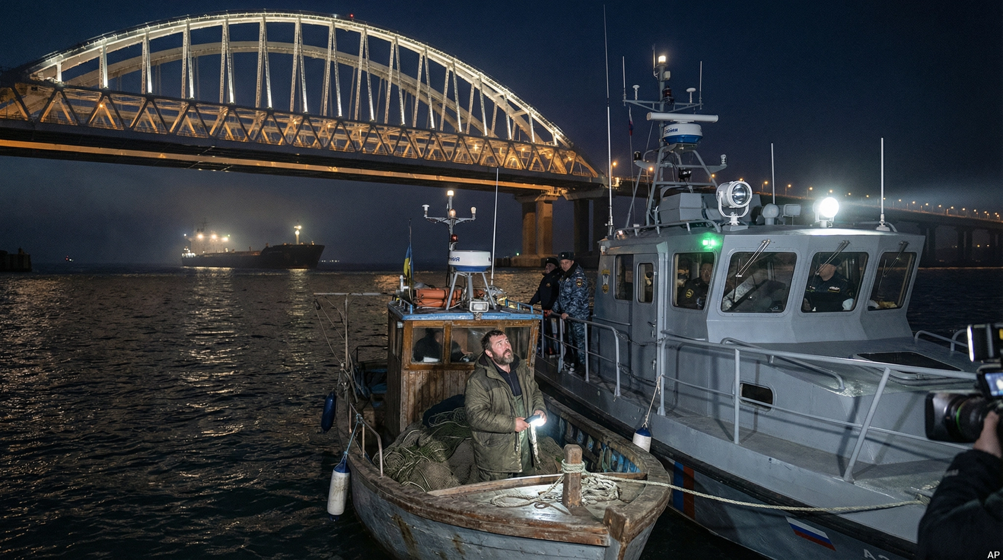 Night scene of the Kerch Strait Bridge, with a Ukrainian fishing boat being inspected by a Russian patrol vessel.