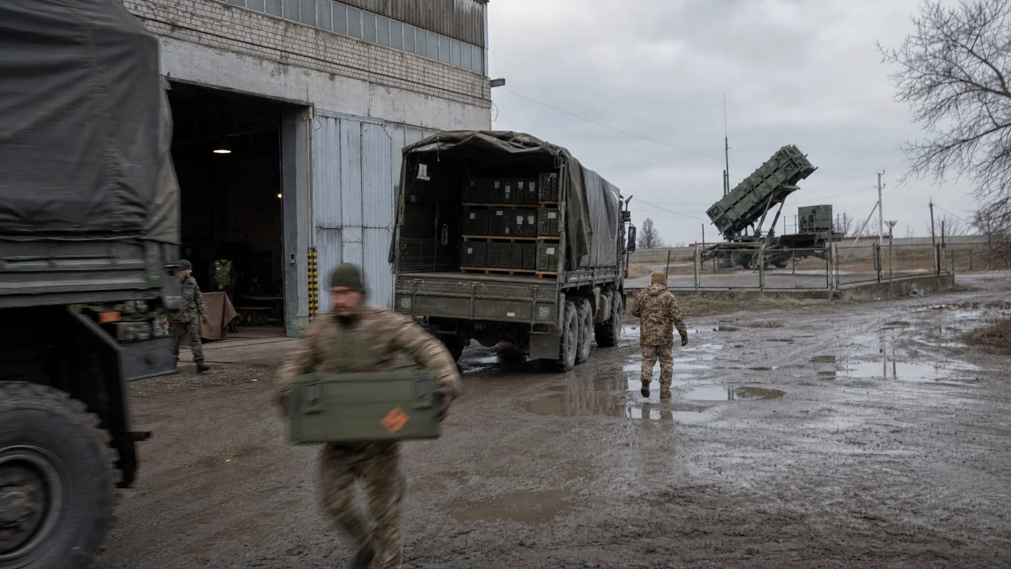 Trucks deliver crates to a rural Ukrainian warehouse as a nearby Patriot missile battery stands ready behind fences. Trucks deliver crates to a rural Ukrainian warehouse as a nearby Patriot missile battery stands ready behind fences.