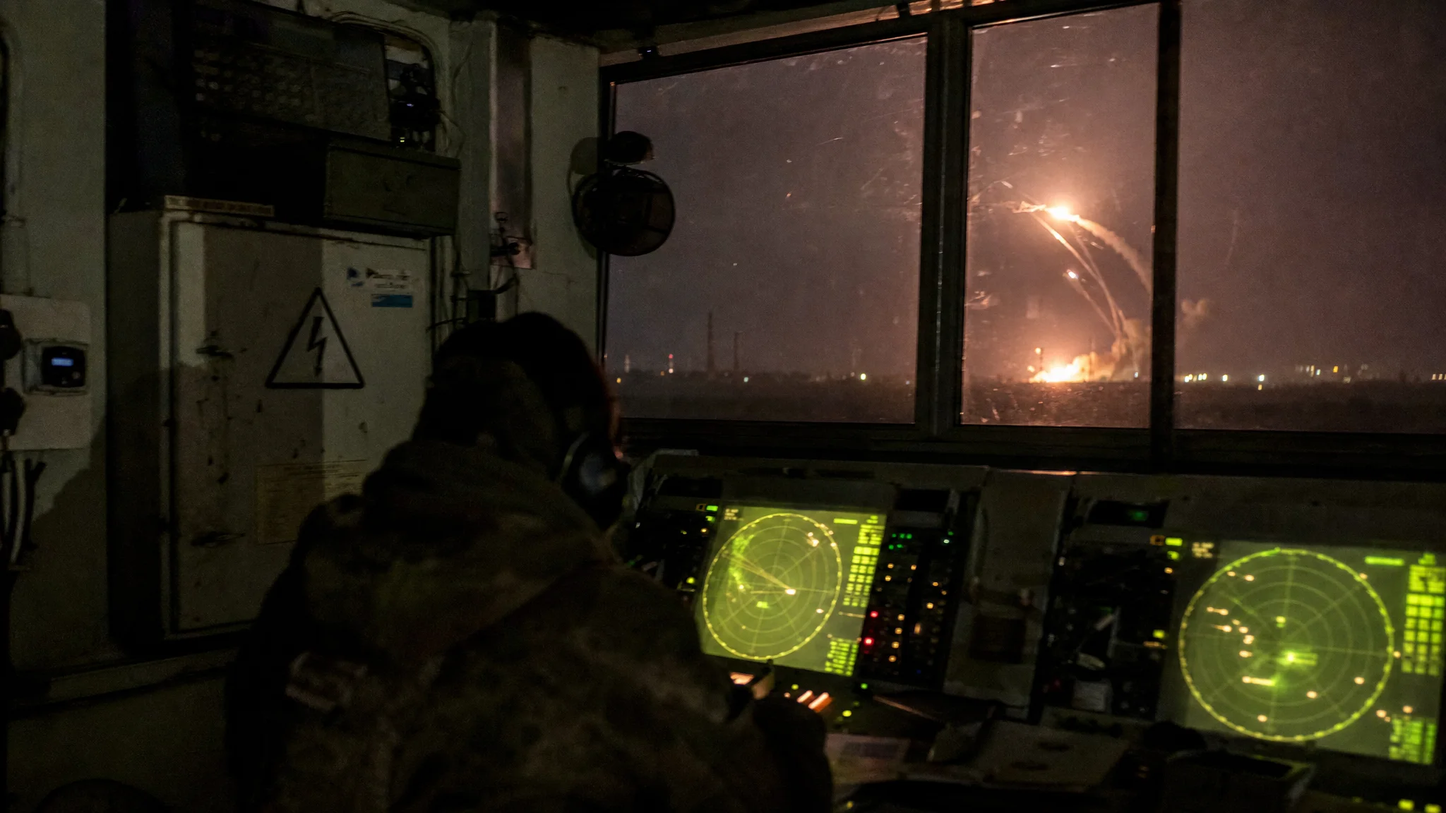 Silhouetted air defense operator watches radar screens; night sky outside is lit by missile interceptors near an industrial site. Silhouetted air defense operator watches radar screens; night sky outside is lit by missile interceptors near an industrial site.