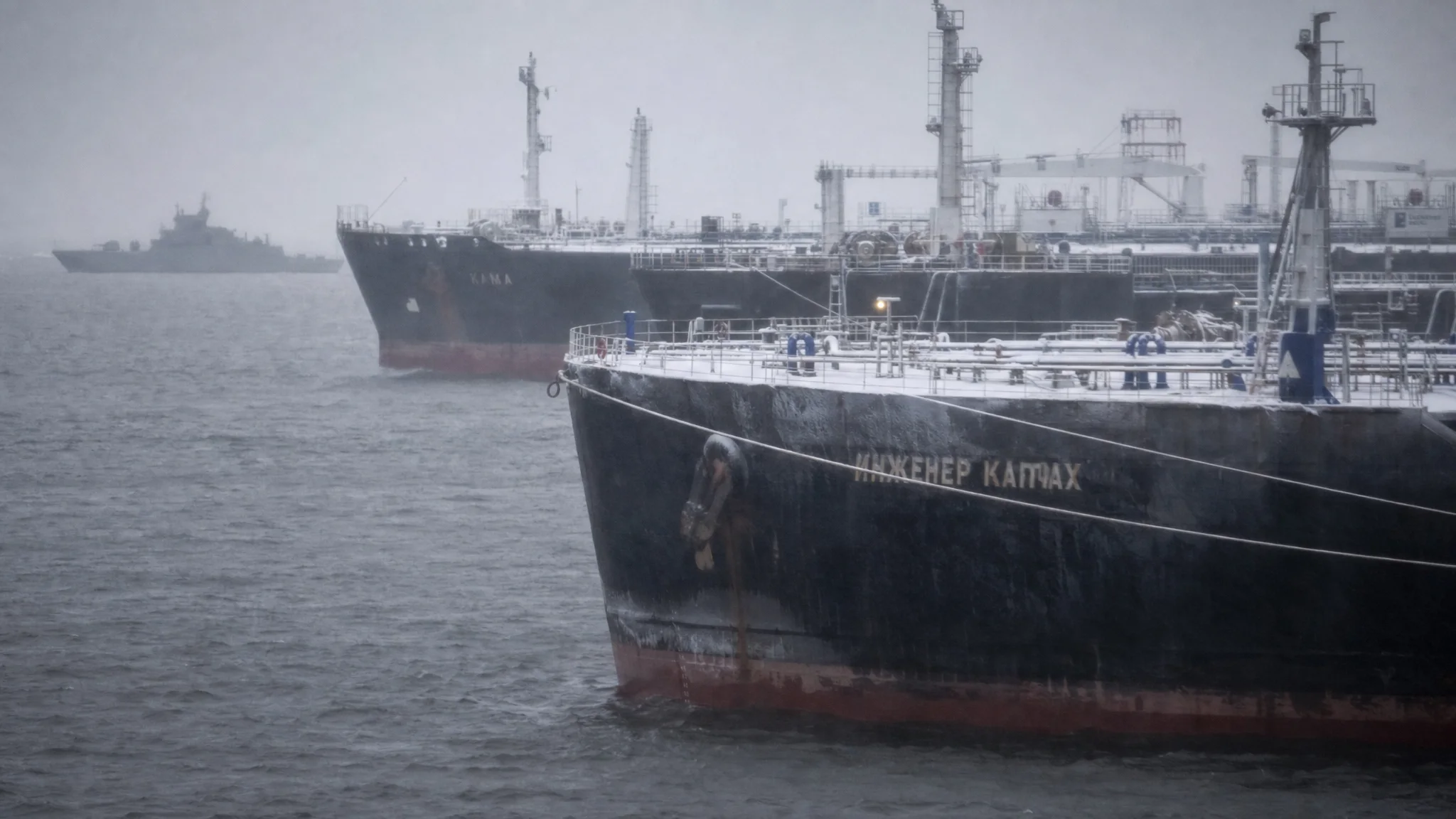 A cluster of stationary, decrepit oil tankers anchored in misty Baltic waters as a navy corvette patrols in the background. A cluster of stationary, decrepit oil tankers anchored in misty Baltic waters as a navy corvette patrols in the background.