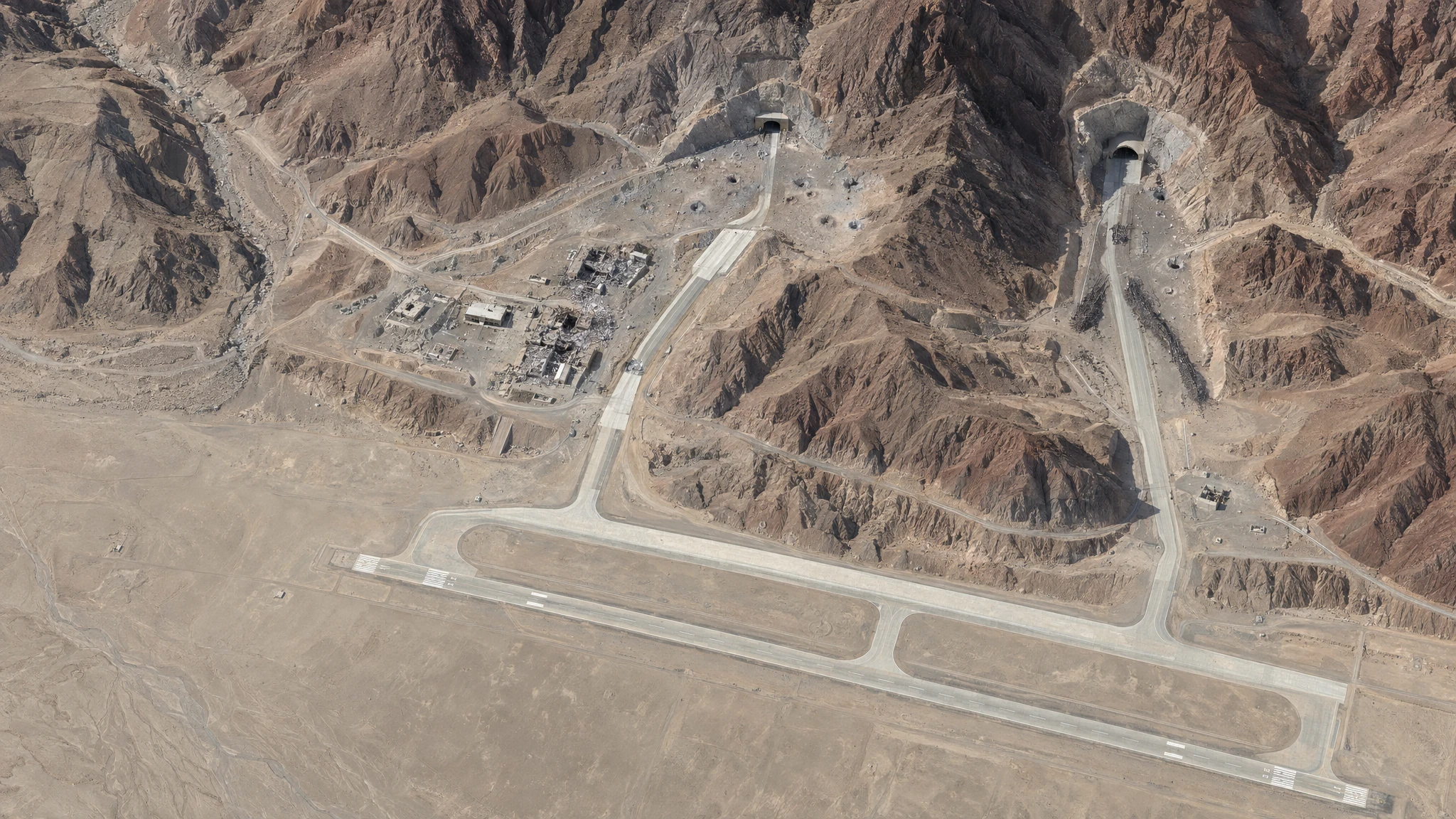 Overhead satellite-style view of a desert airbase showing a cleared runway, taxiways passing through a rocky ridge to mountain tunnel entrances, craters, debris streaks and destroyed buildings. Overhead satellite-style view of a desert airbase showing a cleared runway, taxiways passing through a rocky ridge to mountain tunnel entrances, craters, debris streaks and destroyed buildings.